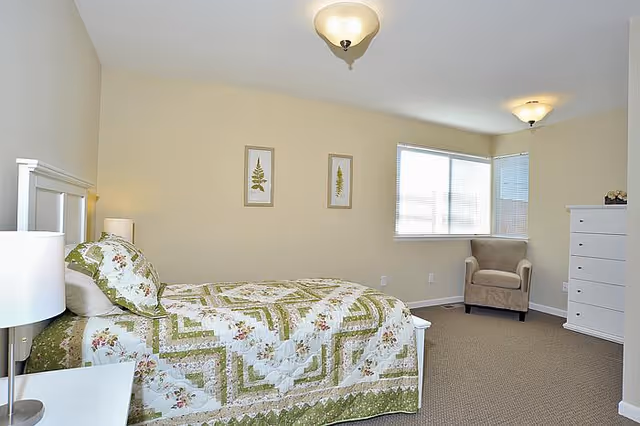 Bright bedroom with a floral green quilted bed, a beige armchair by a corner window, a white dresser, and neutral walls.