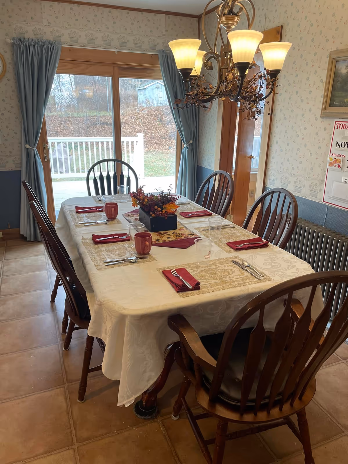 A dining room with a rectangular table set for six, wooden chairs, a chandelier overhead, and sliding glass doors opening to an outdoor deck.