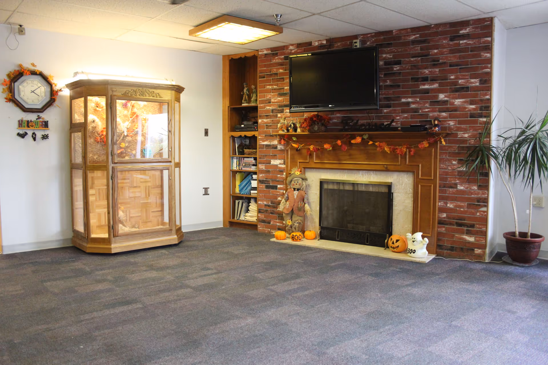 A cozy living room area with a brick fireplace decorated for Halloween, including a scarecrow, pumpkins, and a ghost figure. Above the fireplace is a mounted flat-screen TV. To the left is a wooden display cabinet with glass panels and a wall clock above it. A potted plant is on the right side near the wall.