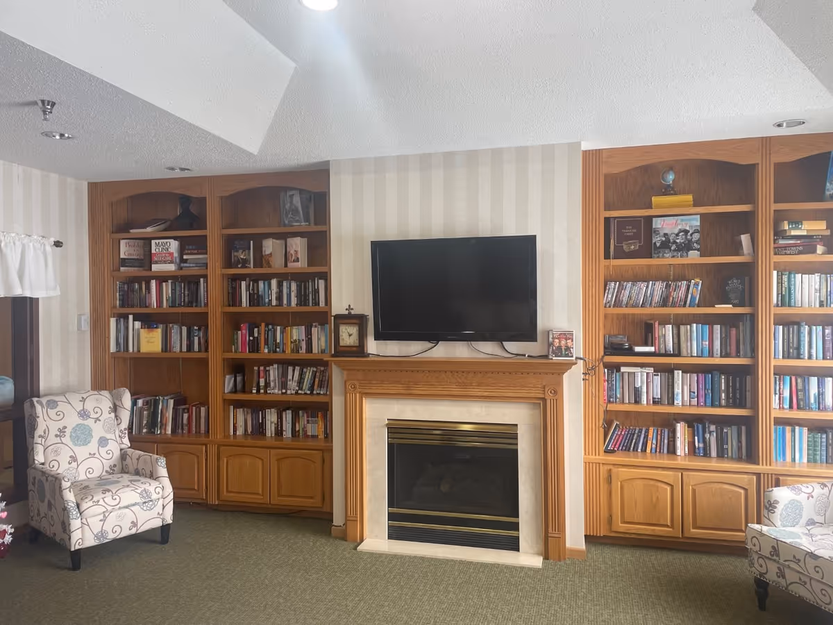 A cozy living room area with built-in wooden bookshelves filled with books on either side of a fireplace. Above the fireplace is a mounted flat-screen TV. Two patterned armchairs are positioned on either side of the room, and the walls have light-colored striped wallpaper.