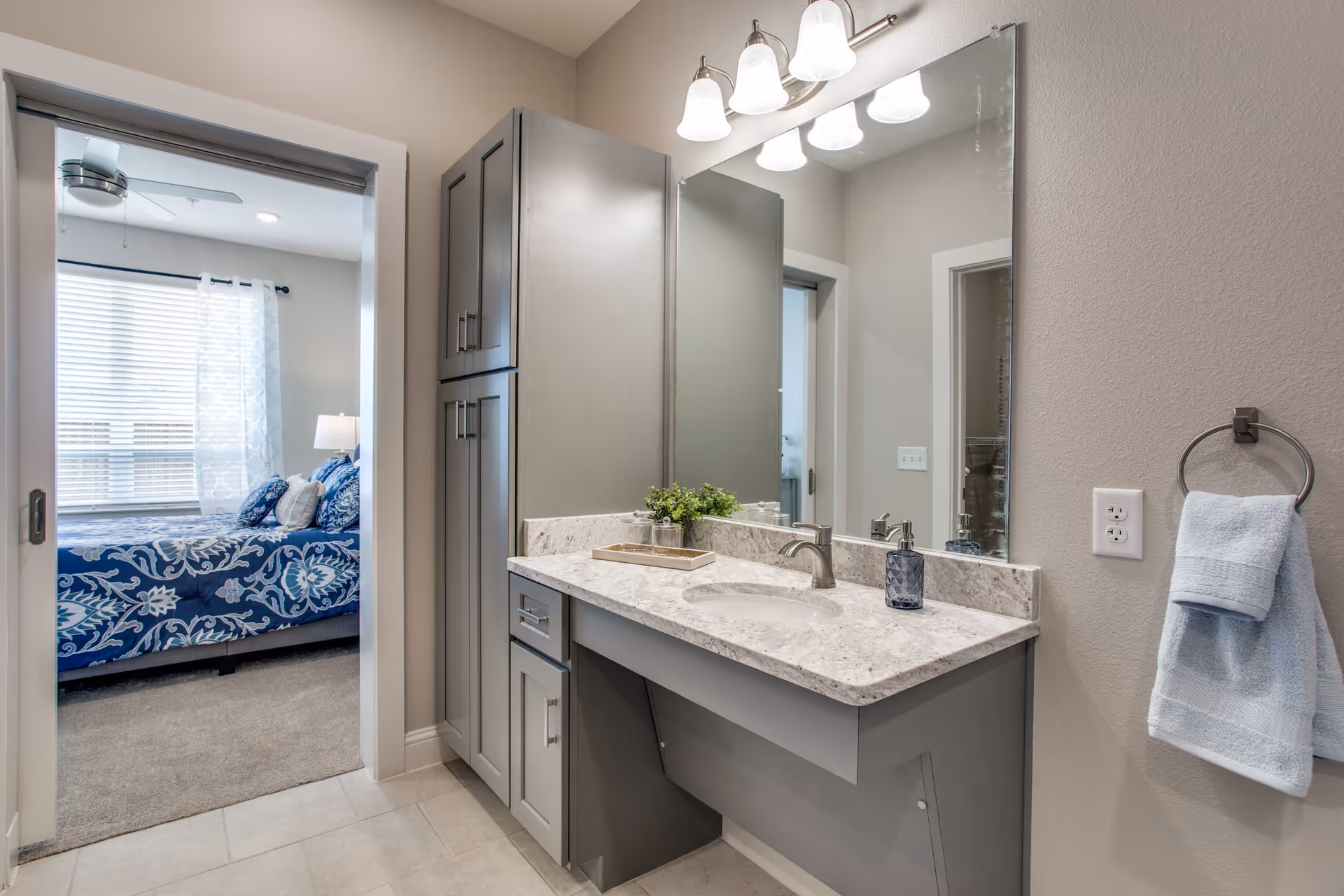 A modern bathroom vanity with a large mirror, granite countertop, and built-in sink. There are three light fixtures above the mirror, a soap dispenser, and a small plant on the counter. To the right, a towel ring holds a folded light blue towel. Through an open doorway, a bedroom with a bed covered in blue and white patterned bedding is visible, along with a window with blinds and sheer curtains.