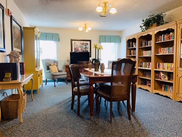 A cozy interior room with a wooden dining table surrounded by six chairs in the center. To the right, there are large wooden bookshelves filled with books. At the back, two windows with light blue valances let in natural light. A comfortable armchair and a TV on a stand are positioned near the windows. The room has a blue patterned carpet and light-colored walls with framed artwork. There are also decorative plants on the table and shelves.