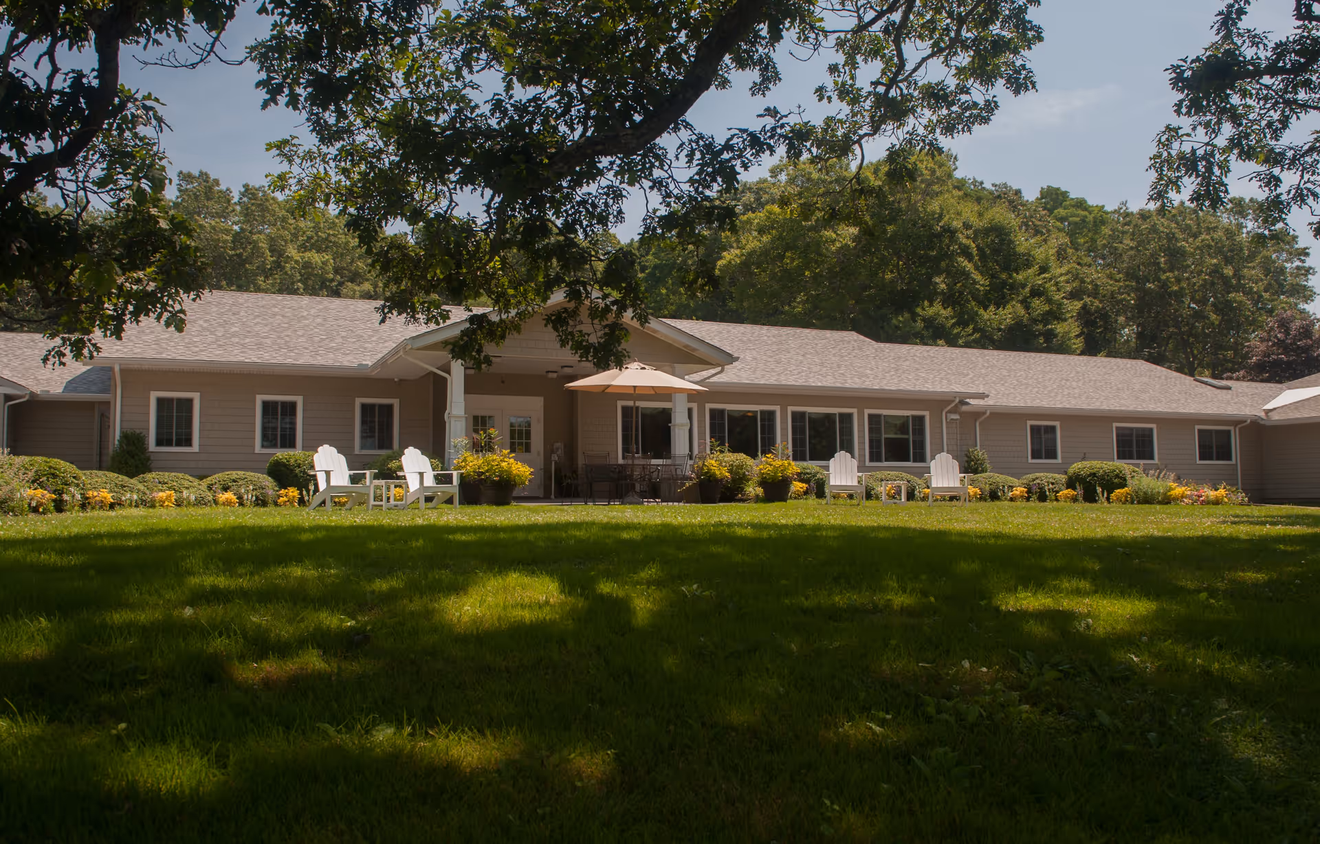 Single-story senior living facility building with a shaded lawn, patio chairs, and an umbrella in front.