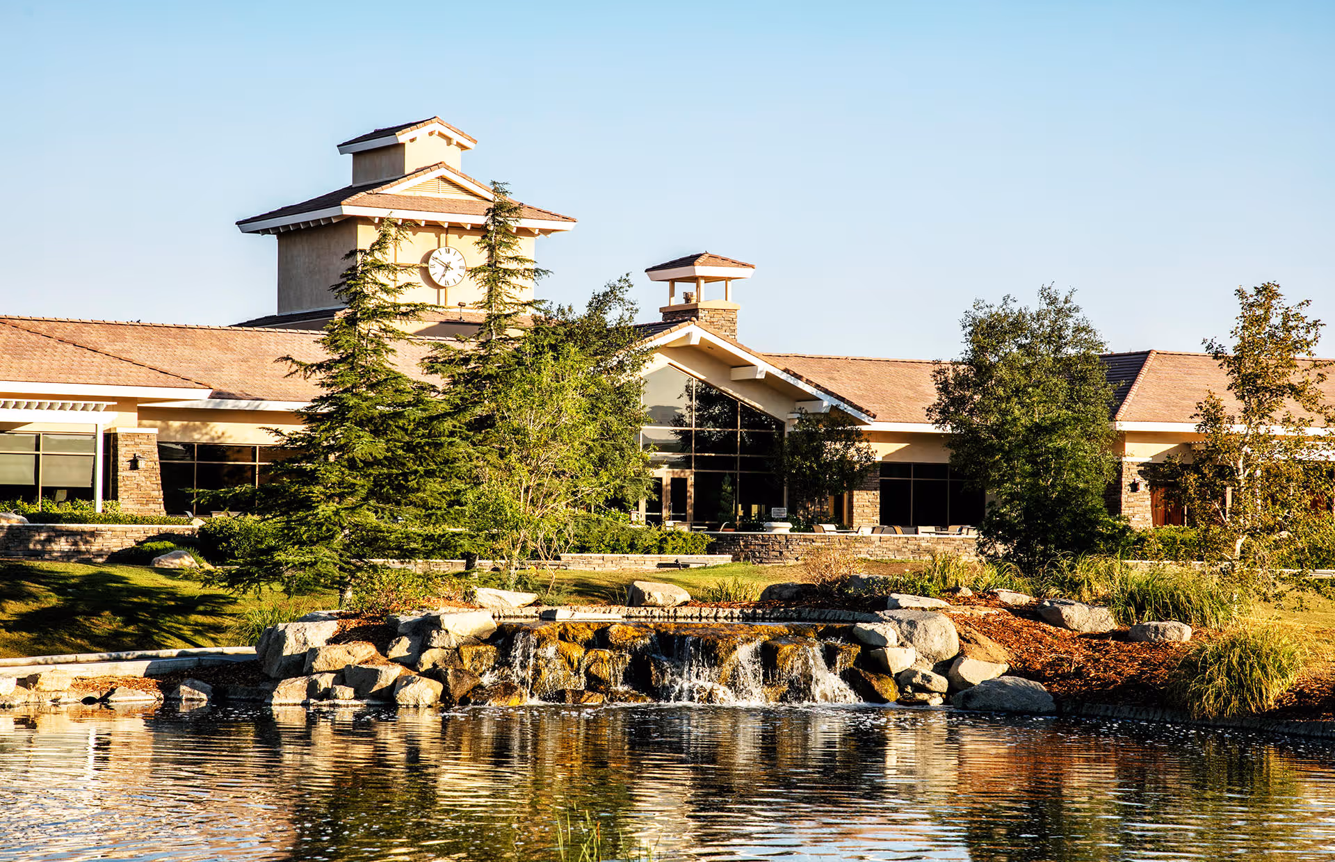 Exterior view of a clubhouse-style building with a clock tower, trees, and a pond with a small waterfall in front.