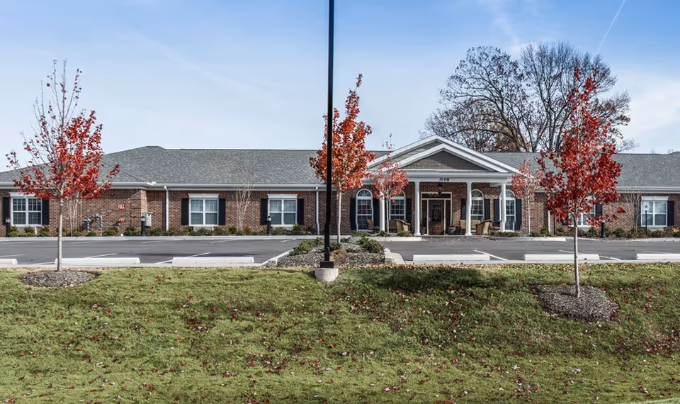 Front exterior view of a single-story brick senior living facility building with a gray shingled roof, white columns at the entrance, several windows with black shutters, and a parking lot in front. There are small trees with red leaves and a grassy area in the foreground under a clear sky.
