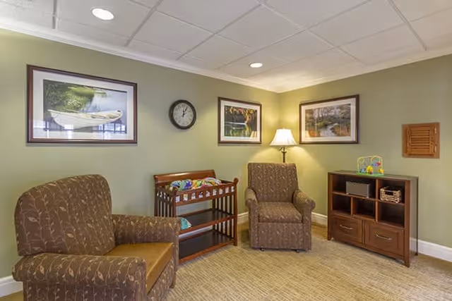 A cozy sitting area with two patterned armchairs, a wooden changing table with baby supplies, a wooden cabinet with cubbies and drawers, a table lamp, a wall clock, and three framed landscape pictures on light green walls.