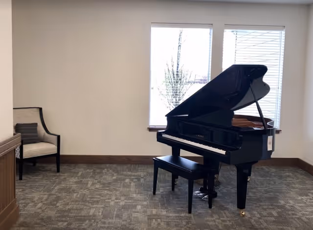 A black grand piano with its lid open sits on a carpeted floor in a room with two large windows covered by white blinds. To the left of the piano, there is a beige upholstered chair with dark wooden legs and a dark pillow. The walls are light-colored with wooden baseboards.