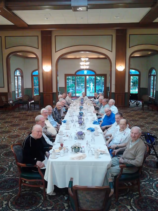 A long dining table set with white tablecloth, cups, glasses, and floral centerpieces in a large room with arched windows and wood paneling. Seated around the table are elderly men, smiling and looking towards the camera.