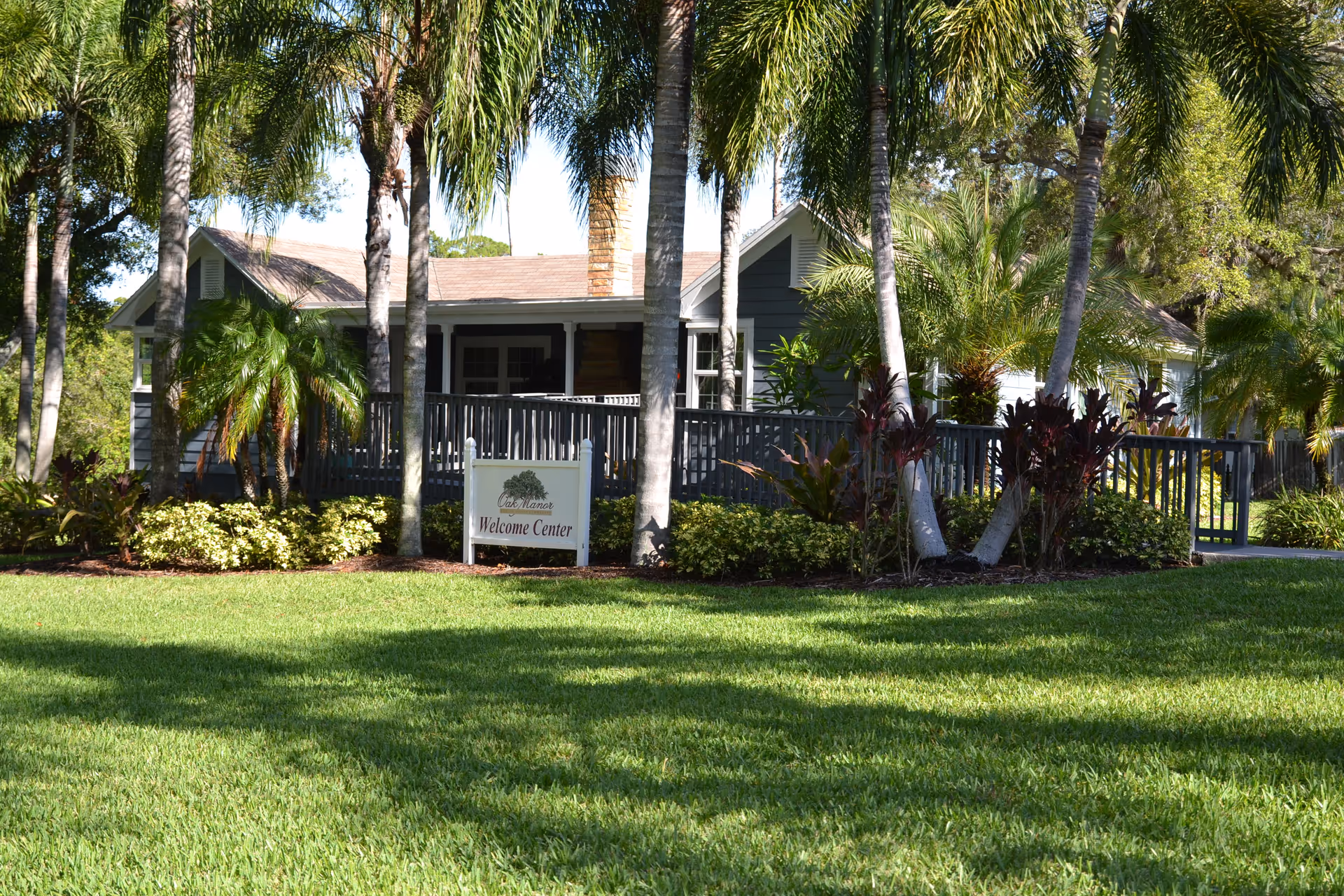 A single-story building surrounded by palm trees and lush greenery with a well-maintained lawn in front. A sign in front of the building reads 'Oak Manor Welcome Center.'