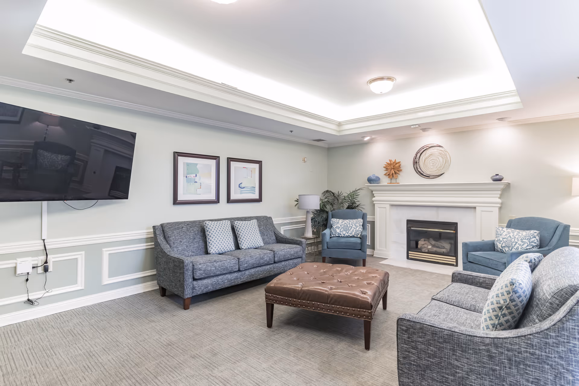 A cozy living room in The Rutherford Assisted Living featuring two gray sofas and two blue armchairs arranged around a large brown tufted ottoman. The room has a white fireplace with decorative items on the mantel, two framed abstract paintings on the wall, a flat-screen TV mounted on the wall, and soft lighting from ceiling fixtures and a table lamp.