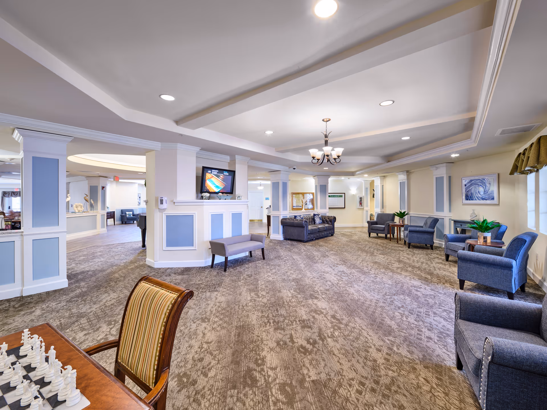 A spacious and well-lit common area in a senior living facility with multiple seating arrangements including sofas and armchairs. The room features a carpeted floor, a ceiling chandelier, and a mounted flat-screen TV above a decorative half wall. There is a chessboard on a table in the foreground and framed artwork on the walls.