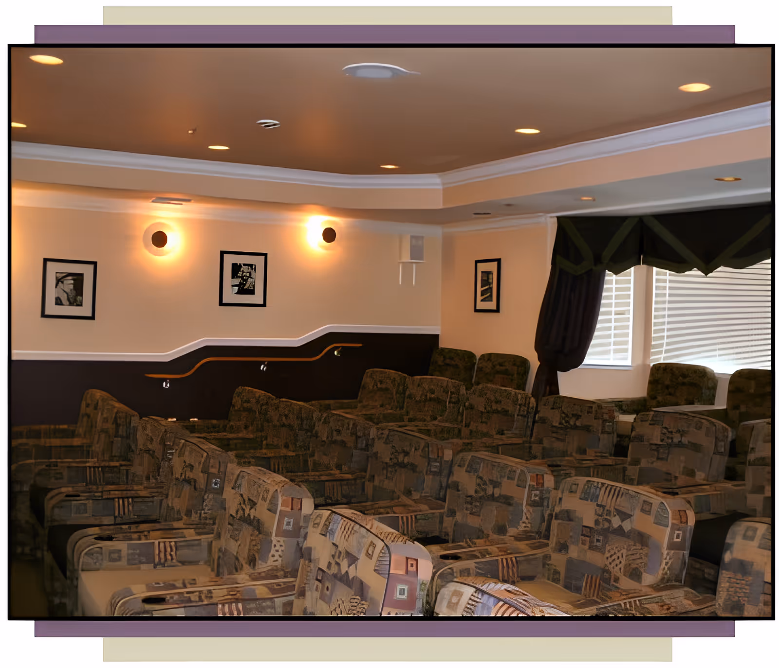 A small communal theater-style room with rows of patterned reclining chairs, wall sconces, framed pictures, and a curtained window.