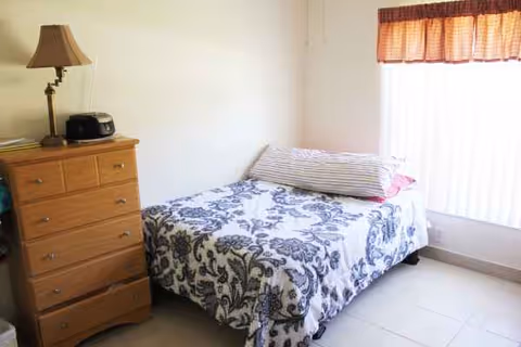 Small bedroom with a single bed, floral bedspread, dresser, lamp, and window blinds.