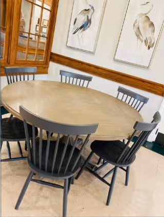 A round wooden dining table surrounded by six black wooden chairs in a room with light-colored walls. Two framed bird paintings hang on the wall above a wooden chair rail, and a wooden-framed glass door is visible in the background.