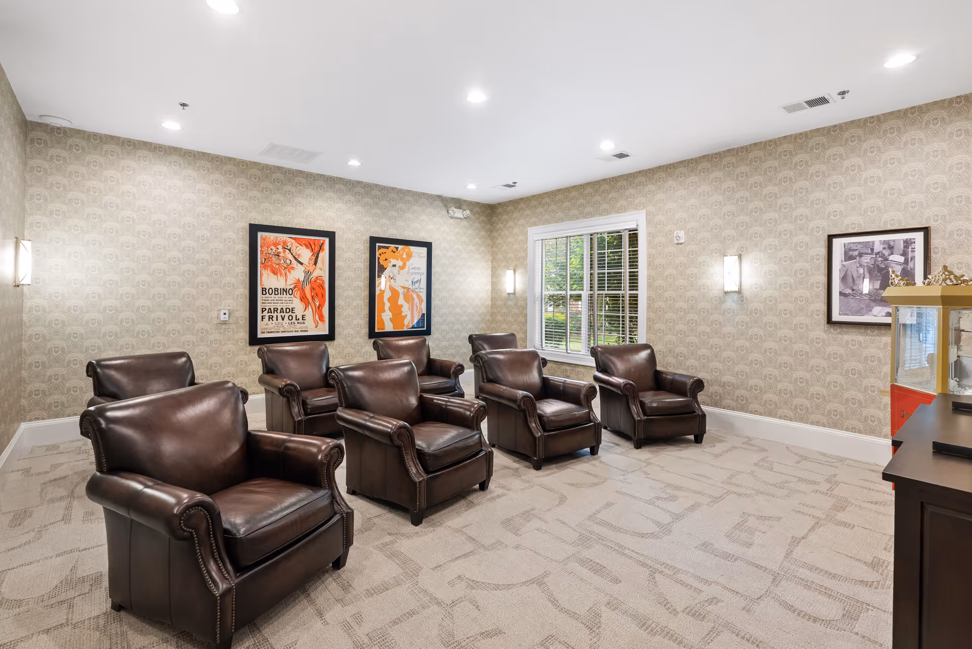 A lounge with six brown leather armchairs arranged in rows facing a window and framed artwork on patterned walls.