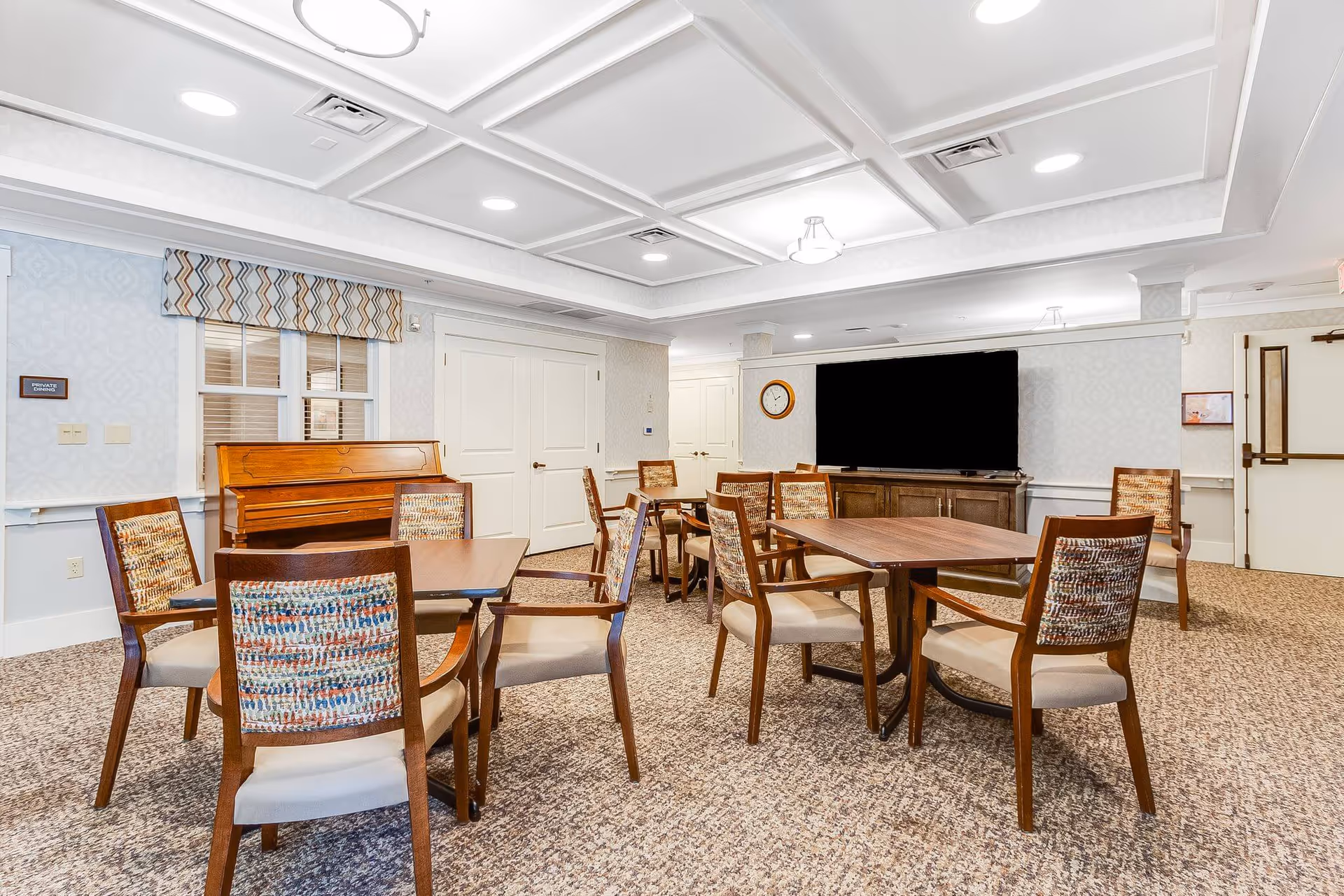 A well-lit room with multiple wooden tables and chairs arranged neatly on a carpeted floor. There is a piano against the wall beneath a window with patterned valances. A large flat-screen TV is mounted on a cabinet on the far wall, with a clock hanging above it. The room has a coffered ceiling with recessed lighting and a door labeled 'Private Dining'.