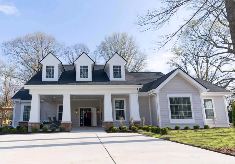 Front exterior view of a single-story building with a dark shingled roof featuring three dormer windows, white columns supporting a covered entrance, and light gray siding. The building is surrounded by a paved driveway and landscaped greenery, with leafless trees in the background under a clear blue sky.