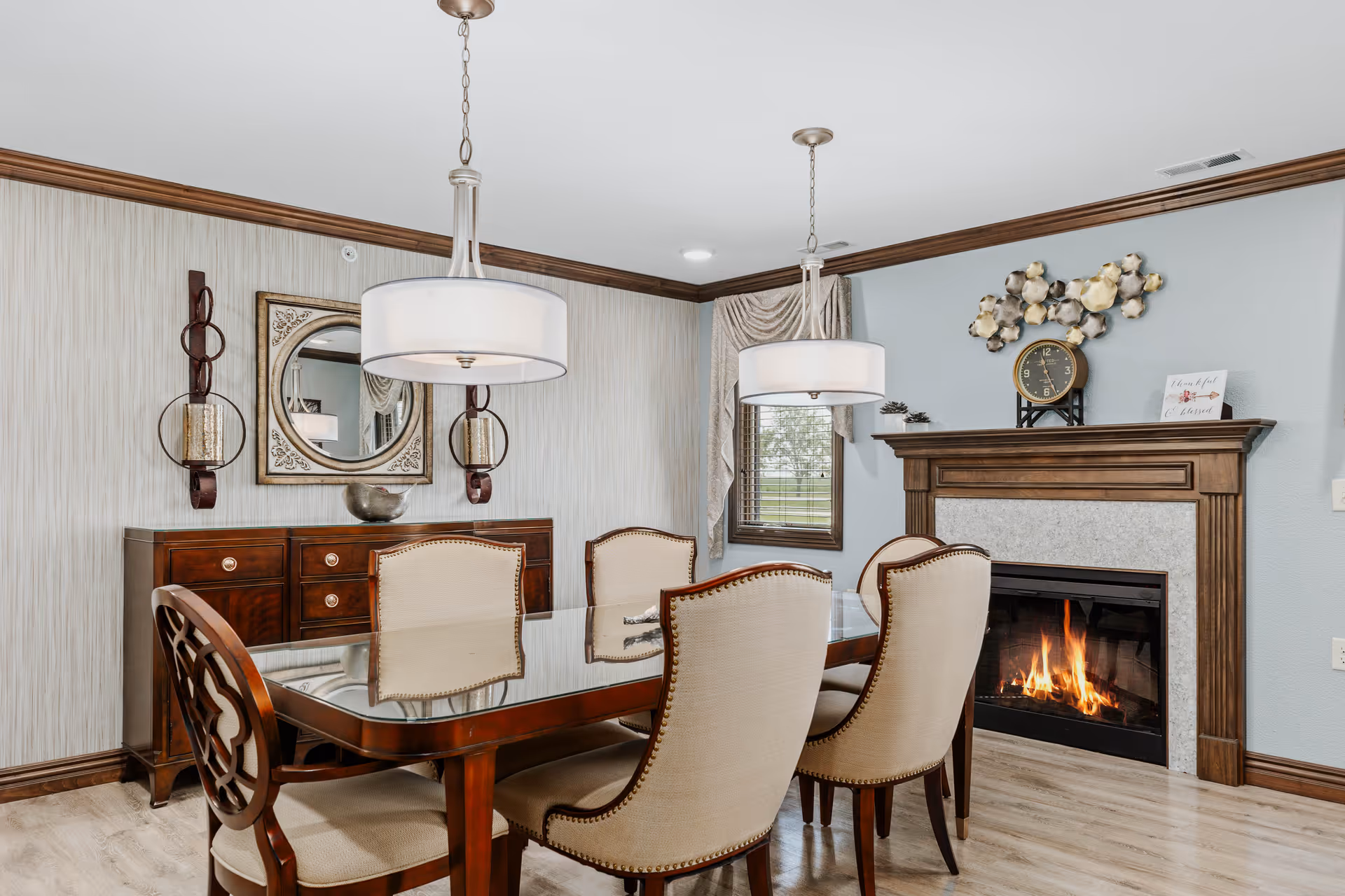A dining room with a wooden table and six upholstered chairs. Two pendant lights hang above the table. There is a wooden sideboard with a decorative mirror and wall sconces on the wall behind it. A fireplace with a wooden mantel and a clock on top is on the right side, with a fire burning inside. A window with curtains is next to the fireplace.