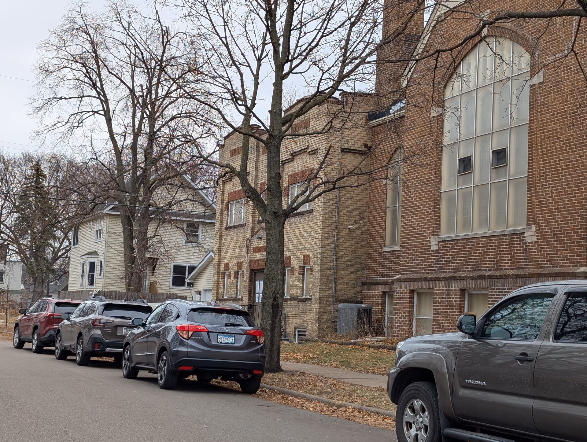 Street view showing several parked cars along a sidewalk with leafless trees and two brick buildings in the background, one with large arched windows.