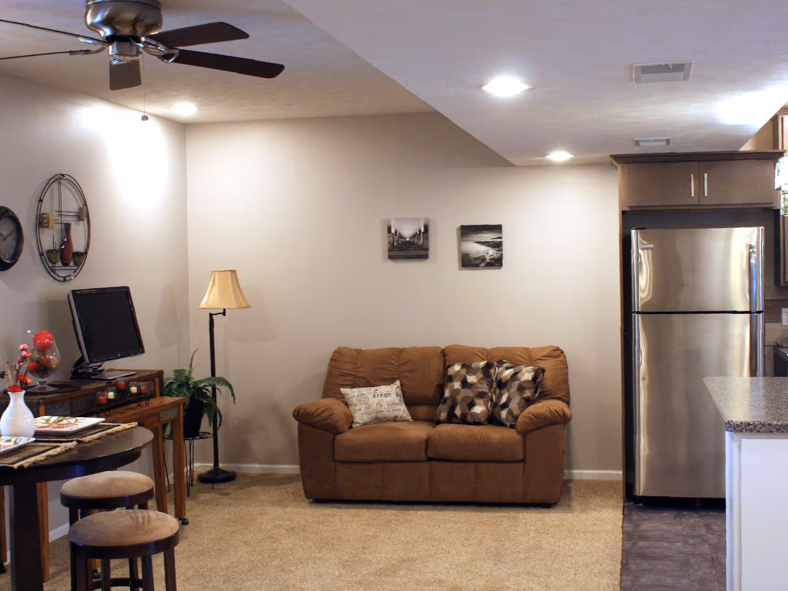 A cozy living area with a brown couch adorned with three decorative pillows, a floor lamp, and two black and white framed pictures on the wall. To the left, there is a small wooden table with a flat-screen TV, a plant, and decorative items. In the foreground, a round dining table with two stools is partially visible. On the right side, a stainless steel refrigerator and part of a kitchen counter with granite countertop are seen. The ceiling has recessed lighting and a ceiling fan with dark blades.