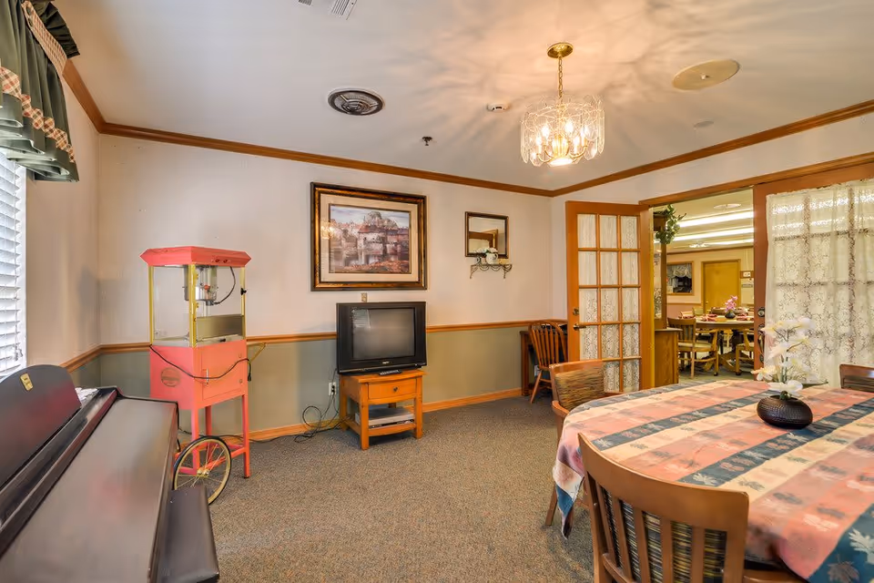 A cozy senior living common area with a piano on the left, a vintage popcorn machine, a small TV on a wooden stand, and a dining table covered with a patterned tablecloth and a flower vase. The room has beige walls with wooden trim, a framed picture, a chandelier, and French doors leading to another room with more tables and chairs.