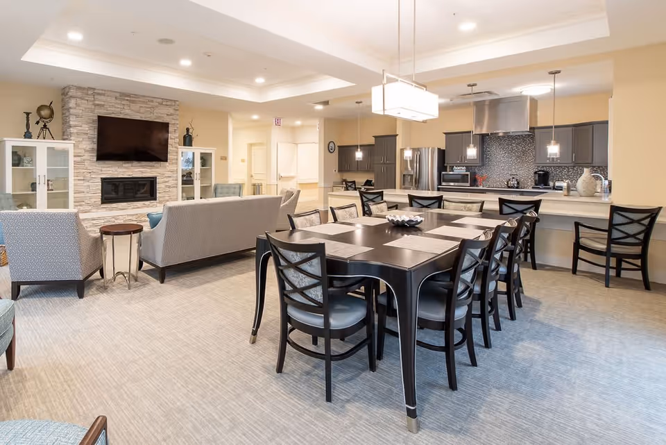 A spacious senior living common area featuring a dining table with eight chairs in the foreground, a modern kitchen with stainless steel appliances and pendant lighting in the background, and a cozy seating area with sofas and armchairs facing a wall-mounted TV above a stone fireplace.