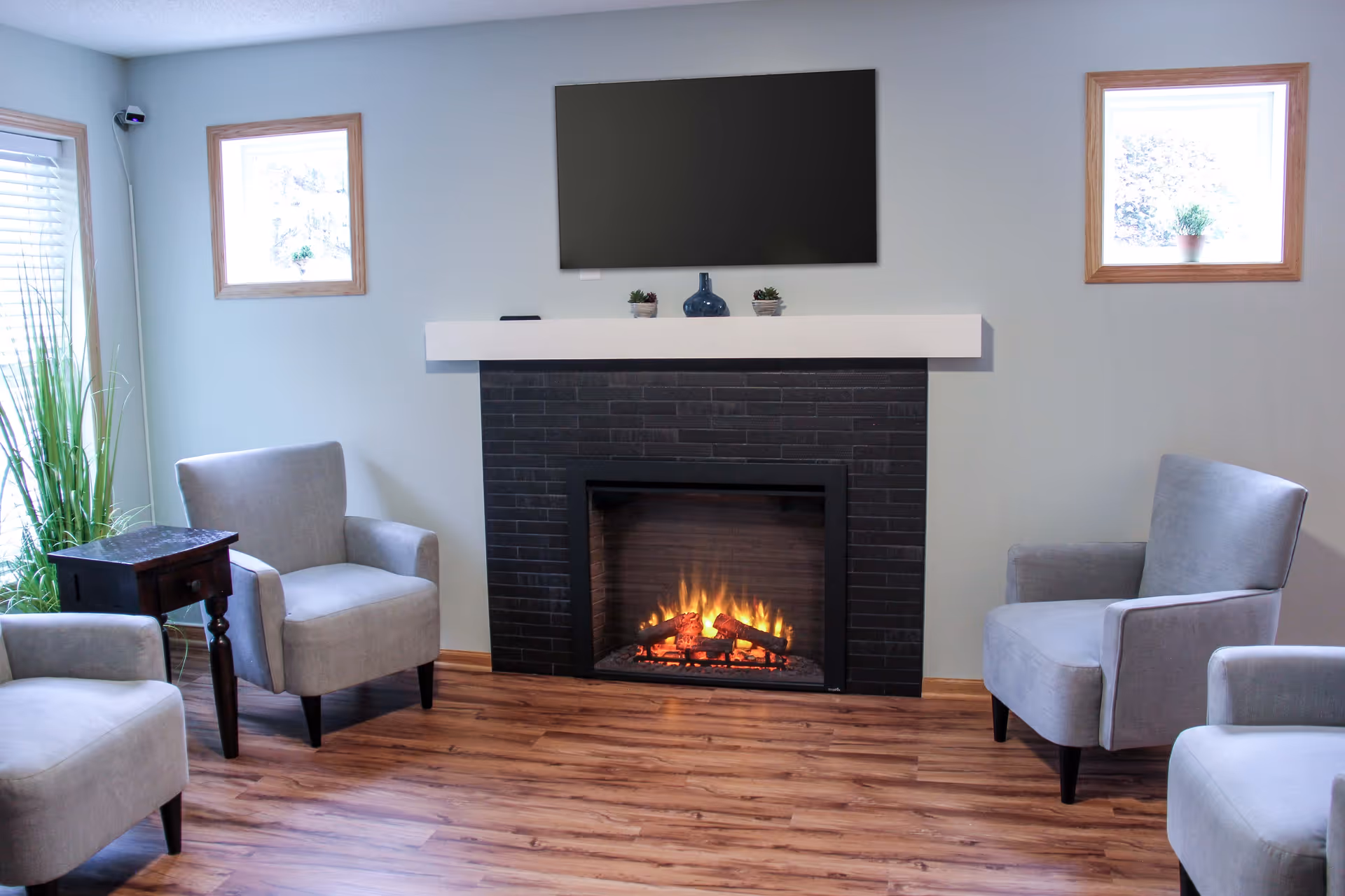 A cozy living room with a modern black fireplace with a white mantel, a flat-screen TV mounted above it, and four gray upholstered armchairs arranged around the fireplace. The room has wooden flooring, two small square windows with wooden frames, and a small wooden side table with a drawer next to one of the chairs. There are small decorative plants on the mantel and a tall green plant near a window with blinds.