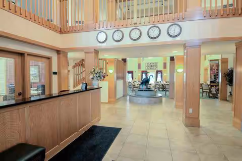 Interior view of a senior living facility lobby with a wooden reception desk on the left, a tiled floor, and a central water fountain. Above the entrance to a dining area, there are six clocks showing different time zones. The space features warm lighting, wooden railings on the upper level, and seating areas visible in the background.