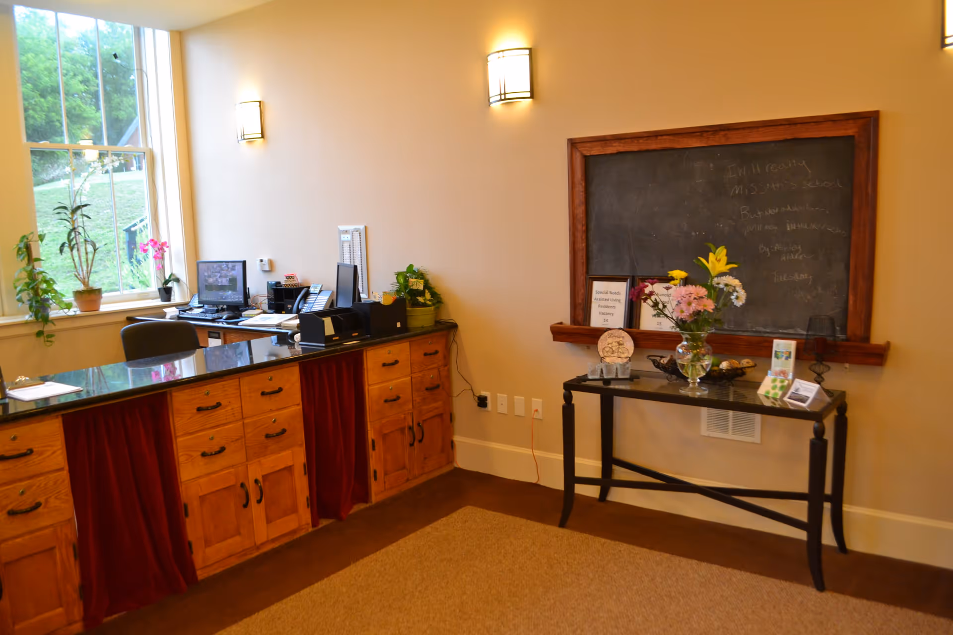 Reception area with a wooden desk featuring a black countertop, office chair, computer, and various office supplies. A large window with potted plants on the sill lets in natural light. On the right wall, there is a blackboard with some writing and a small table below it holding a vase with flowers and informational materials.