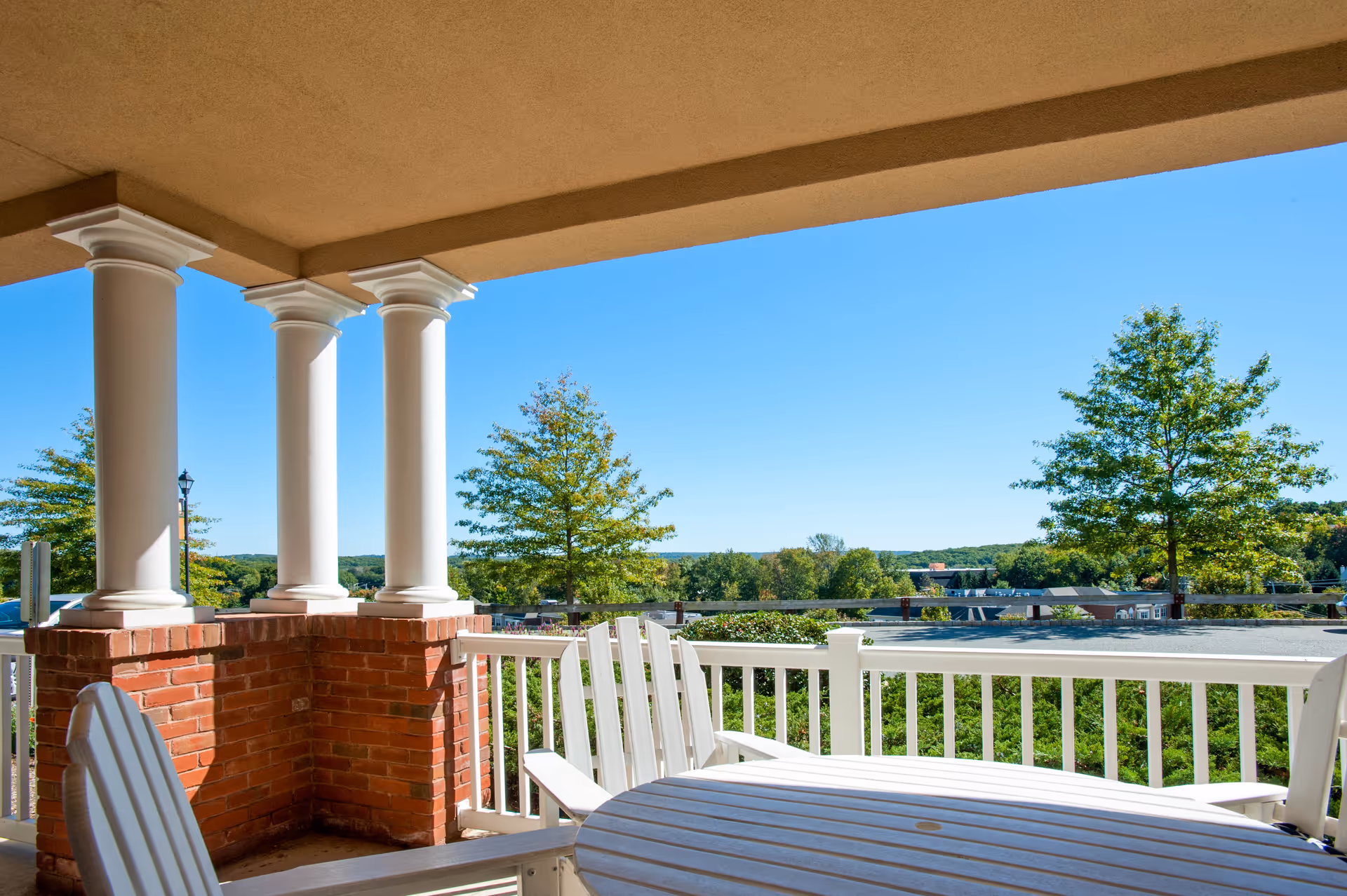 Covered balcony with white columns, railing and patio furniture overlooking trees and a distant landscape.