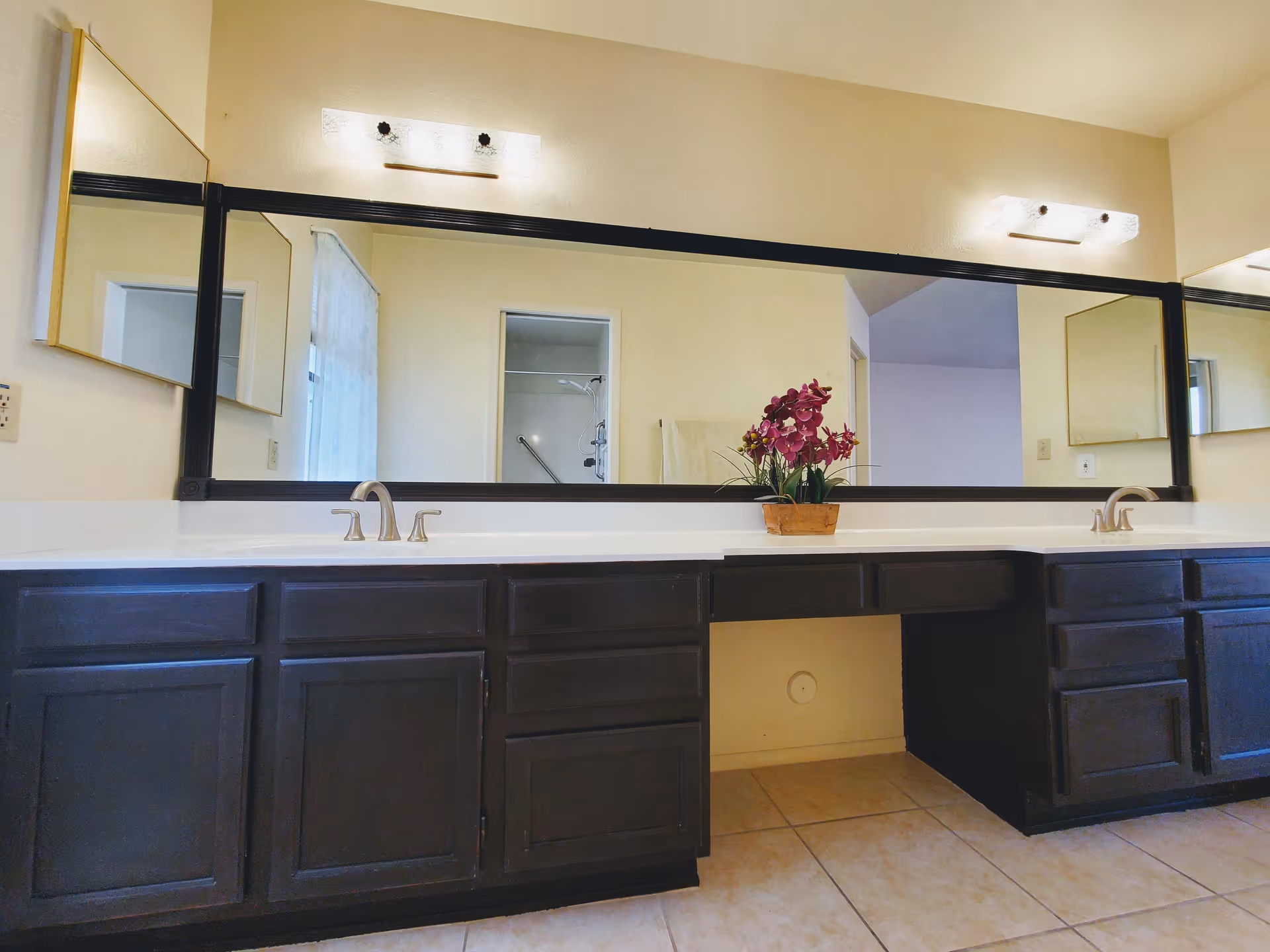 A bathroom vanity with two sinks and dark wood cabinets beneath a large rectangular mirror. There are two light fixtures above the mirror, and a small potted plant with purple flowers is placed on the countertop. The reflection in the mirror shows a doorway leading to a shower area and part of an adjacent room.