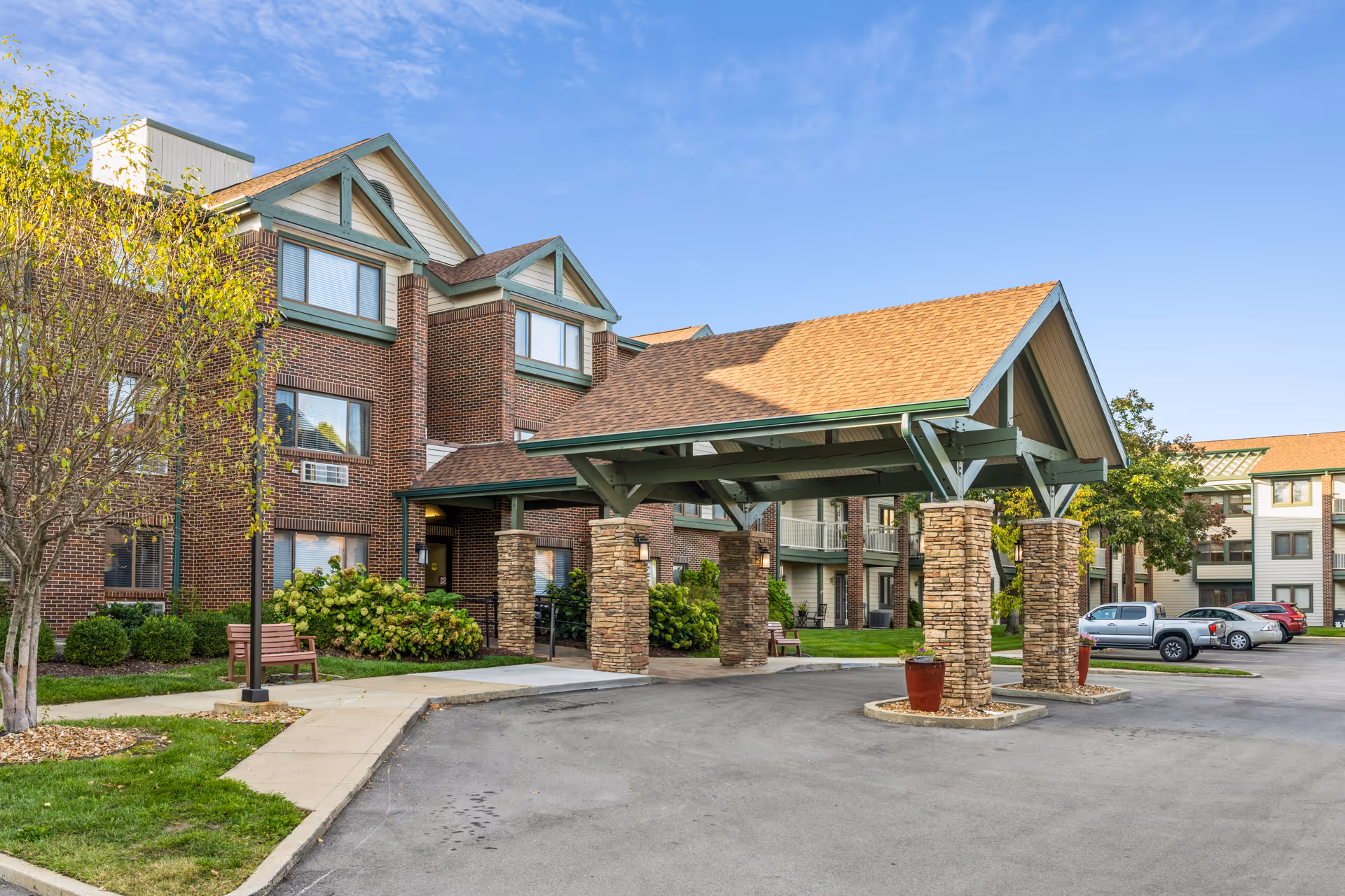Front entrance of a brick senior living building with a covered porte-cochere, parking area, and landscaping.