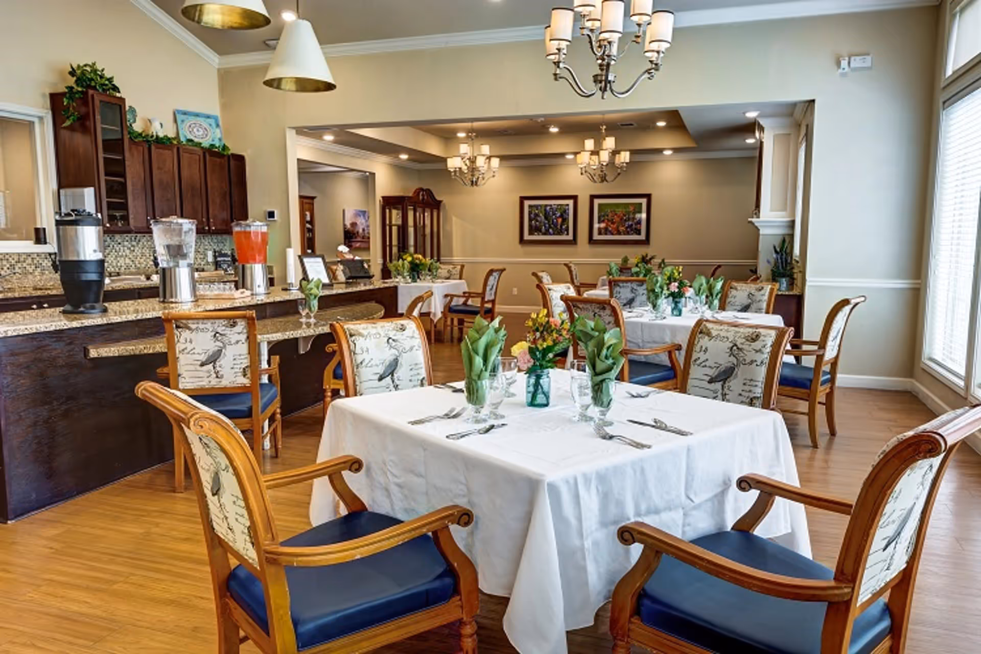 Bright, formal dining room with tables set with white tablecloths, wooden chairs, floral centerpieces and a beverage service counter in the background.