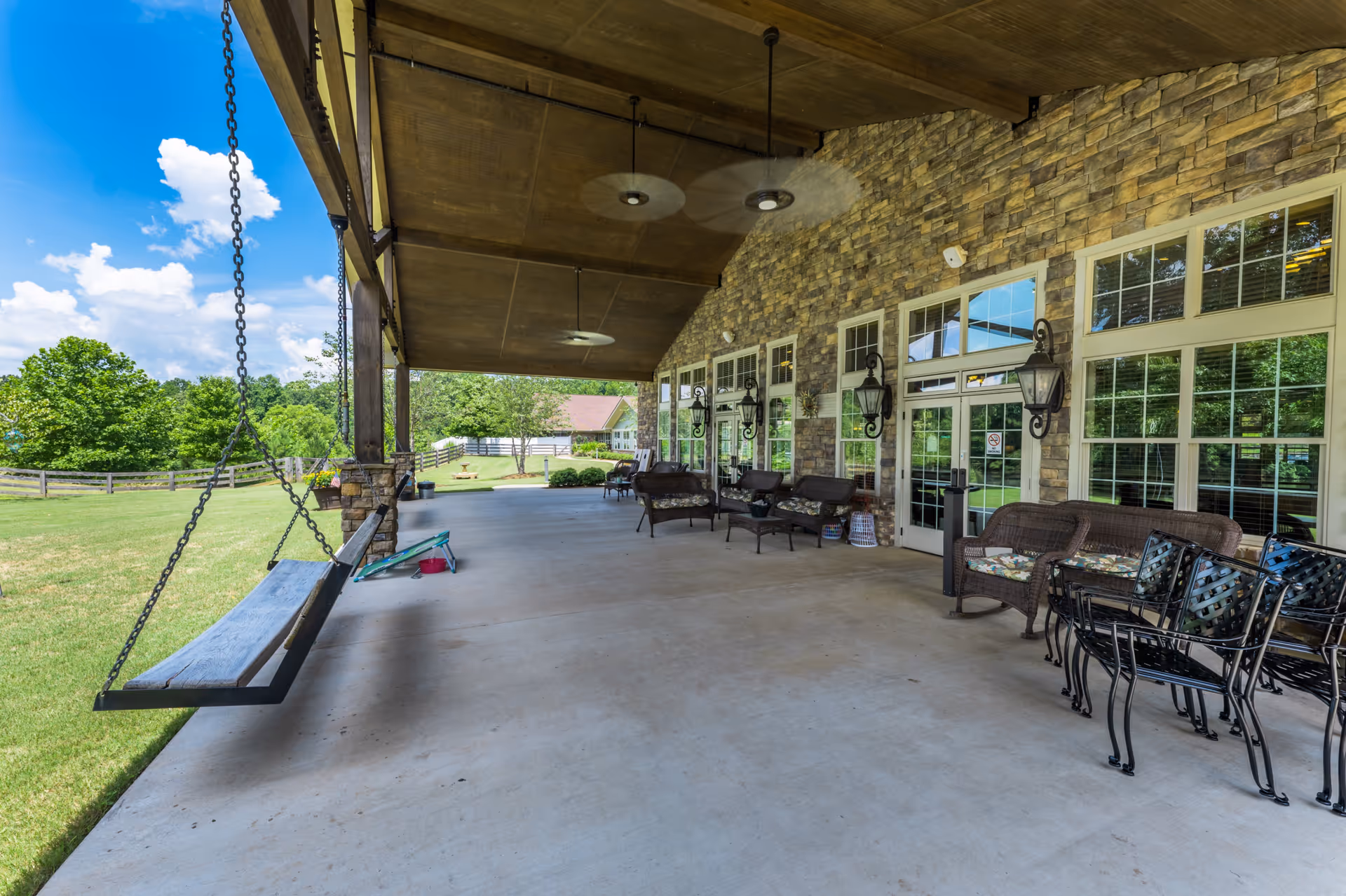 Covered outdoor patio area with a wooden swing hanging from the ceiling on the left side, several cushioned chairs and benches along the stone wall on the right, large windows and glass doors, ceiling fans, and a view of a grassy lawn and trees under a blue sky with clouds.