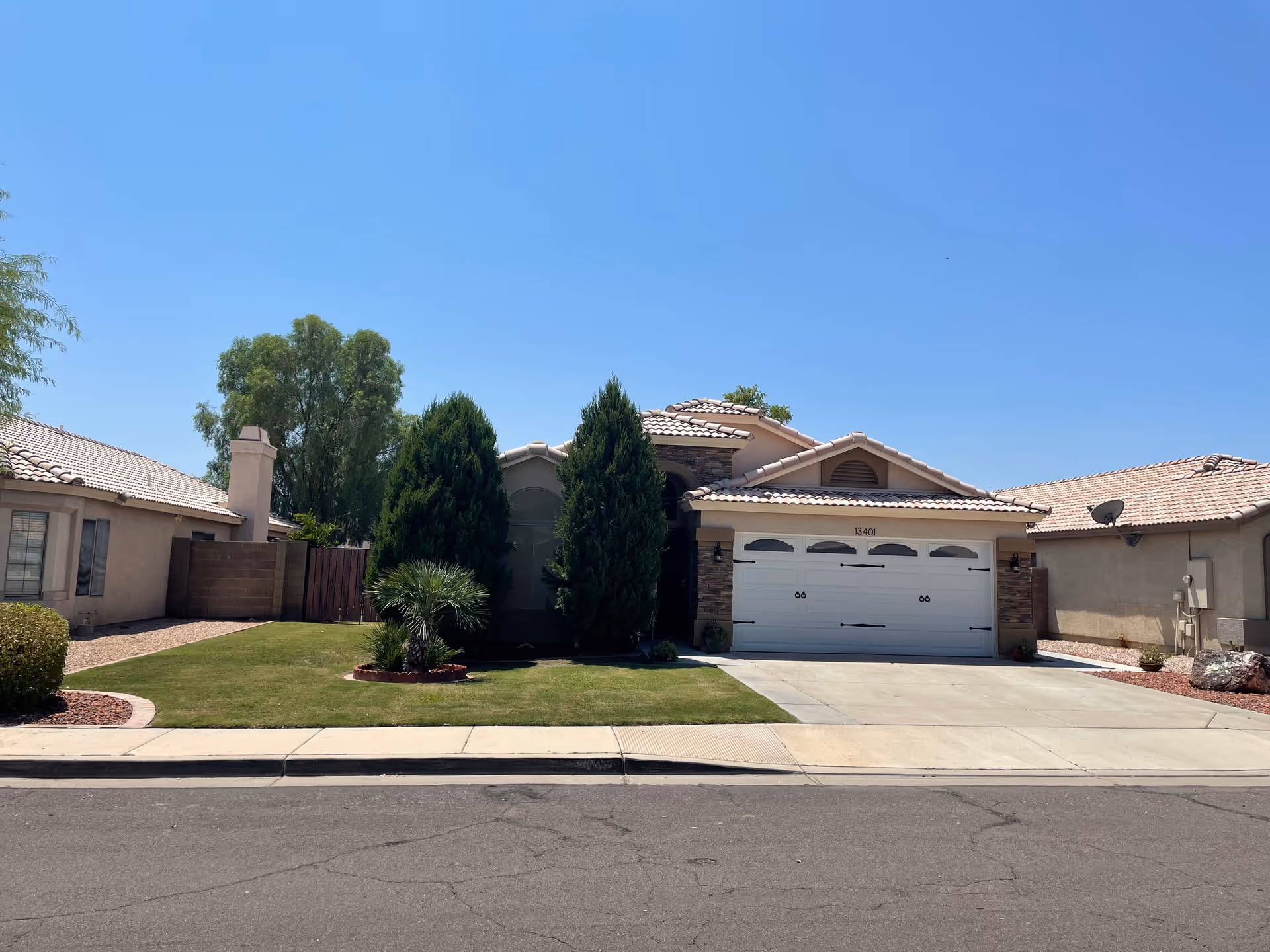 Front exterior view of a single-story house with a tiled roof, a white garage door, and a well-maintained lawn with trees and shrubs under a clear blue sky.