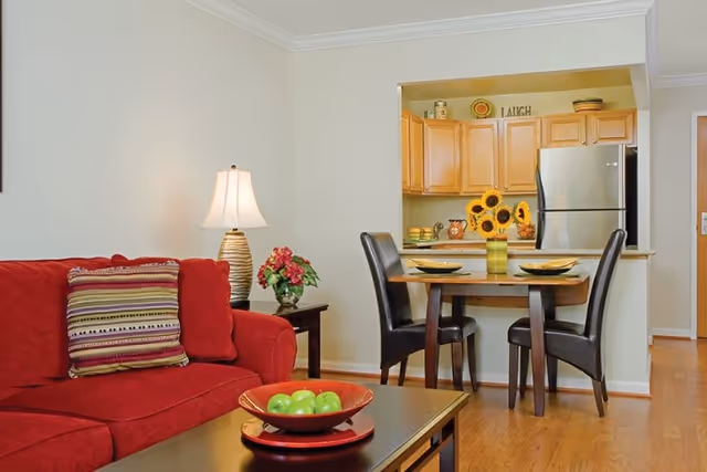 A cozy living and dining area featuring a red sofa with a striped pillow, a side table with a lamp and a flower arrangement, a wooden coffee table with a bowl of green apples, and a small dining table set with two black chairs and a vase of sunflowers. The background shows a kitchen with wooden cabinets and a stainless steel refrigerator.