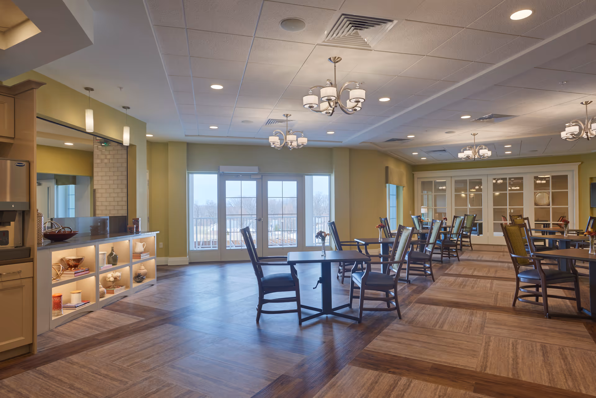 Spacious dining area with multiple round tables and chairs, a serving counter with lit display shelves, large windows, and chandeliers.