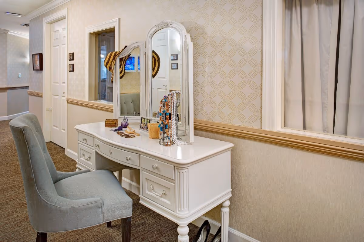 A white vanity table with a tri-fold mirror and jewelry on top, accompanied by a gray upholstered chair in a decorated hallway.