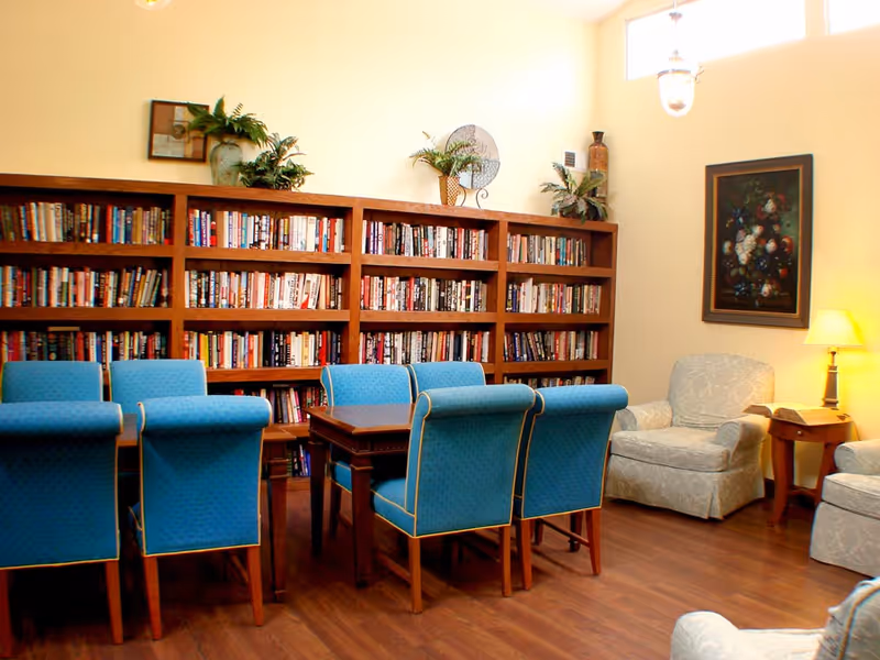 A cozy reading room with a wooden bookshelf filled with books along the wall, a wooden table surrounded by six blue upholstered chairs, and two light-colored armchairs near a small wooden side table with a lamp. The room has wooden flooring, light yellow walls, a framed floral painting, and decorative plants on top of the bookshelf.