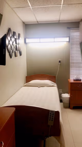 A small nursing home bedroom with a single bed made with white sheets and a pillow. The bed has a wooden headboard and footboard, and a remote control attached to the footboard. On the right side of the bed is a wooden nightstand with a box of tissues on top. A window with blinds is partially visible behind the nightstand. The wall above the bed has decorative metal wall art, and there is a bright horizontal light fixture above the headboard. The floor is tiled, and a small trash can is next to the nightstand.