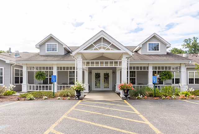 Front exterior view of a single-story senior living facility building with a covered entrance, two handicap parking signs, flower pots with colorful flowers, and a clear sky with some clouds.