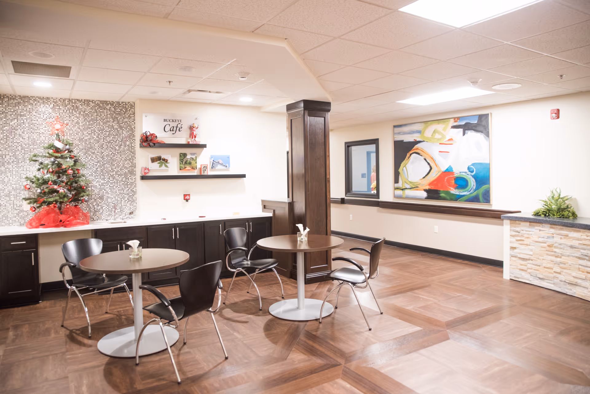Interior view of a small cafe area named Buckeye Cafe with two round tables and black chairs. There is a decorated Christmas tree on a counter with dark cabinets below, a mosaic tile backsplash, and some small decorative items on shelves. The floor has a wood pattern, and there is a colorful abstract painting on the wall.