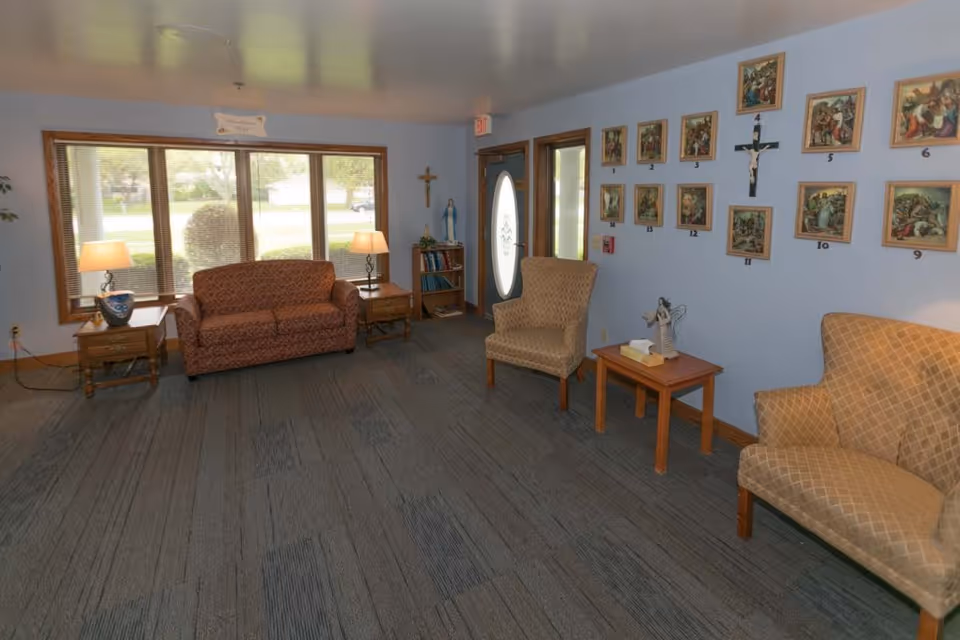 A cozy assisted living facility common area with a patterned sofa, two armchairs, two side tables with lamps, a bookshelf with religious statues, and a wall decorated with framed religious artwork and a crucifix. Large windows let in natural light and a door with an oval glass panel is visible.
