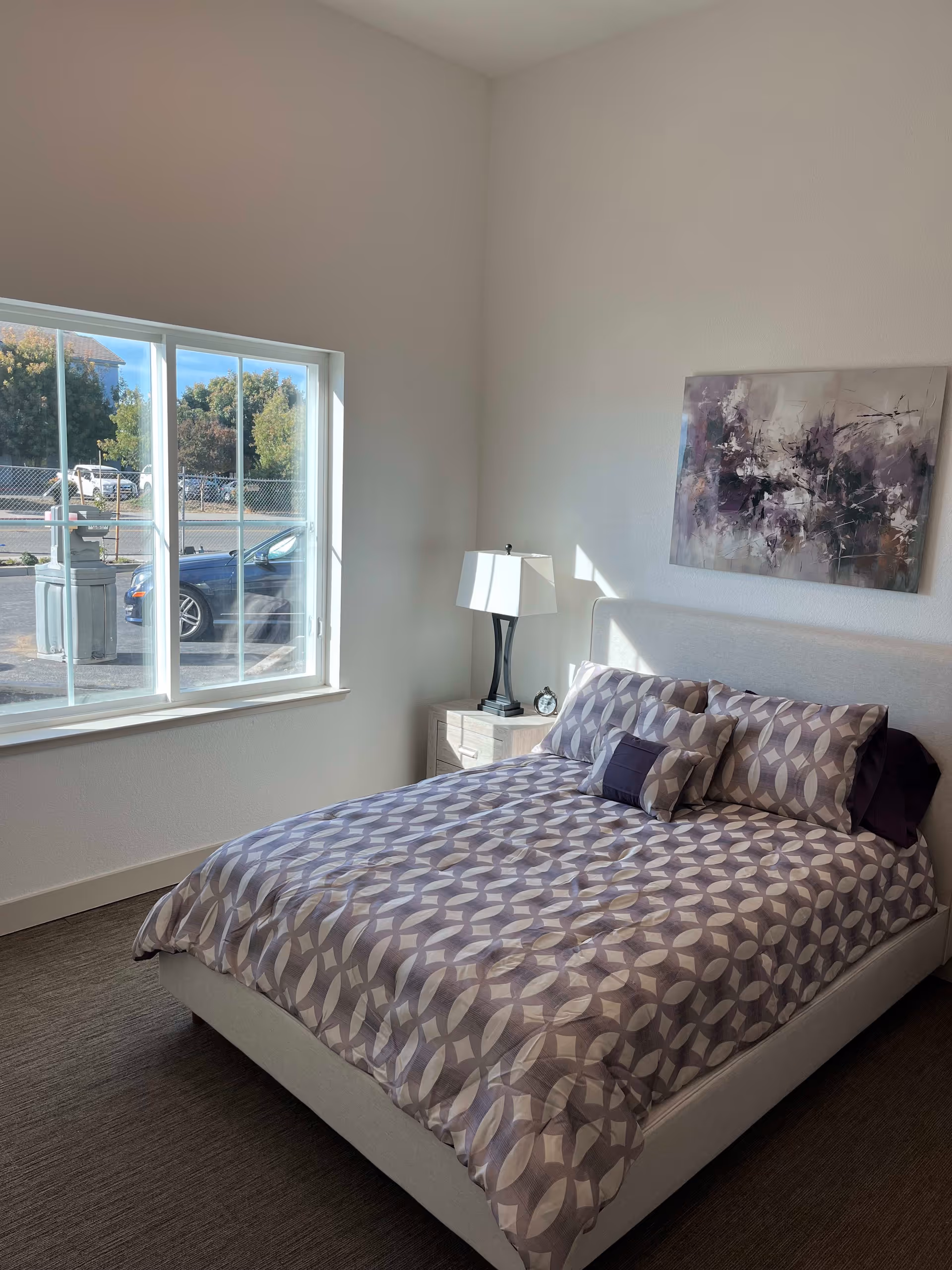 A bright bedroom with a patterned bed, nightstand and lamp, abstract wall art, and a large window looking out to the street.