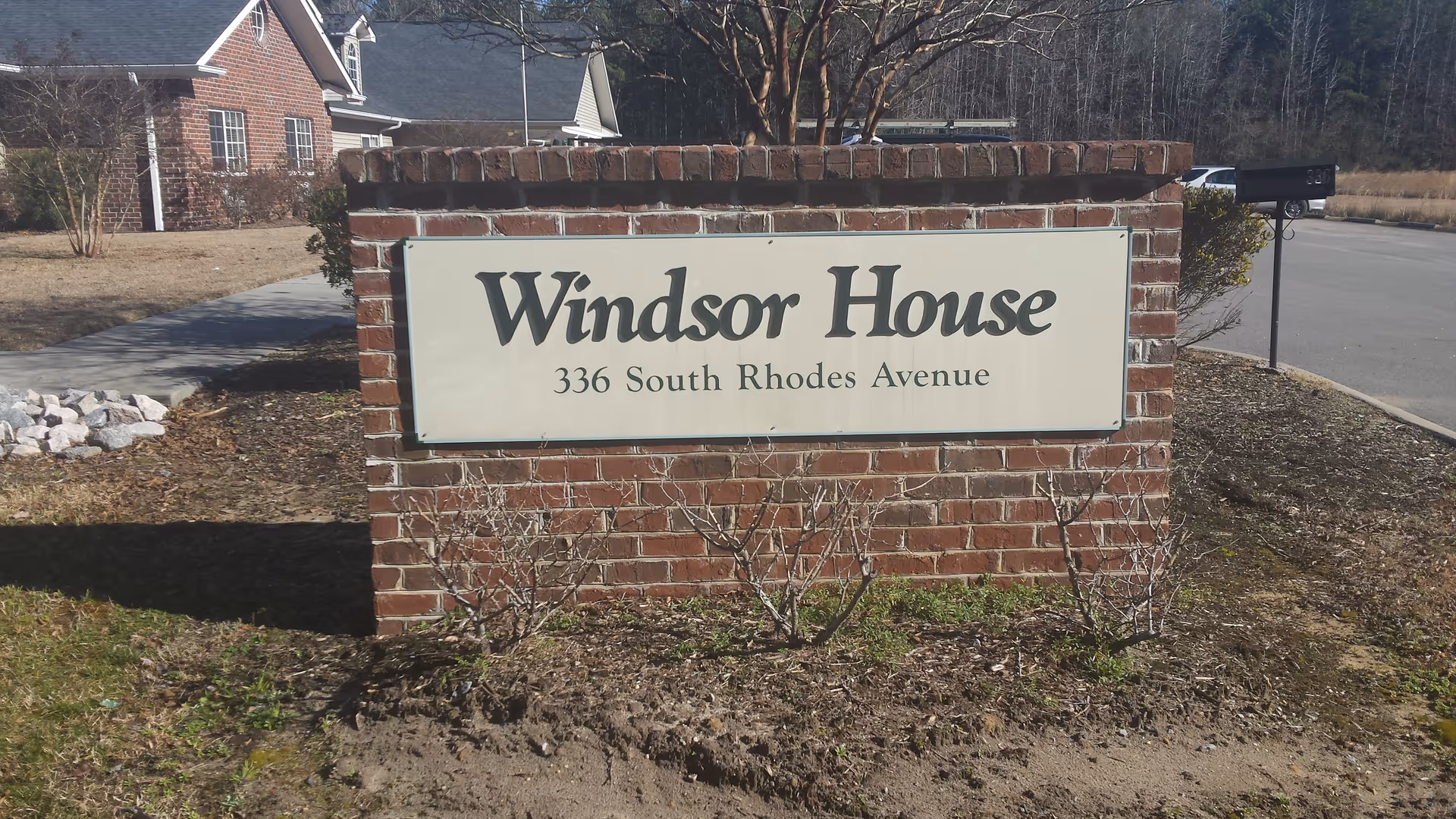 A brick signpost with a white rectangular sign that reads 'Windsor House 336 South Rhodes Avenue' situated on a grassy area near a sidewalk and a road. In the background, there is a brick building and some leafless trees.