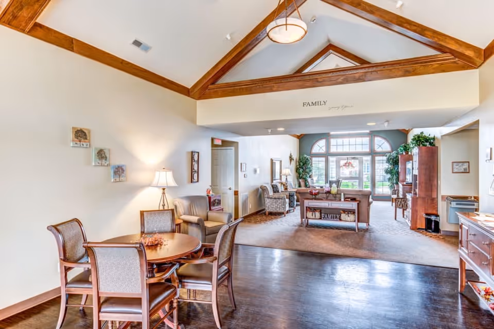 A bright and spacious living room area with vaulted ceilings featuring wooden beams. The room includes a round wooden table with four chairs, a floor lamp, comfortable armchairs, and a sofa. Large windows at the far end allow natural light to fill the space, and there are decorative plants and framed pictures on the walls. The word 'FAMILY' is displayed above the entrance to the seating area.