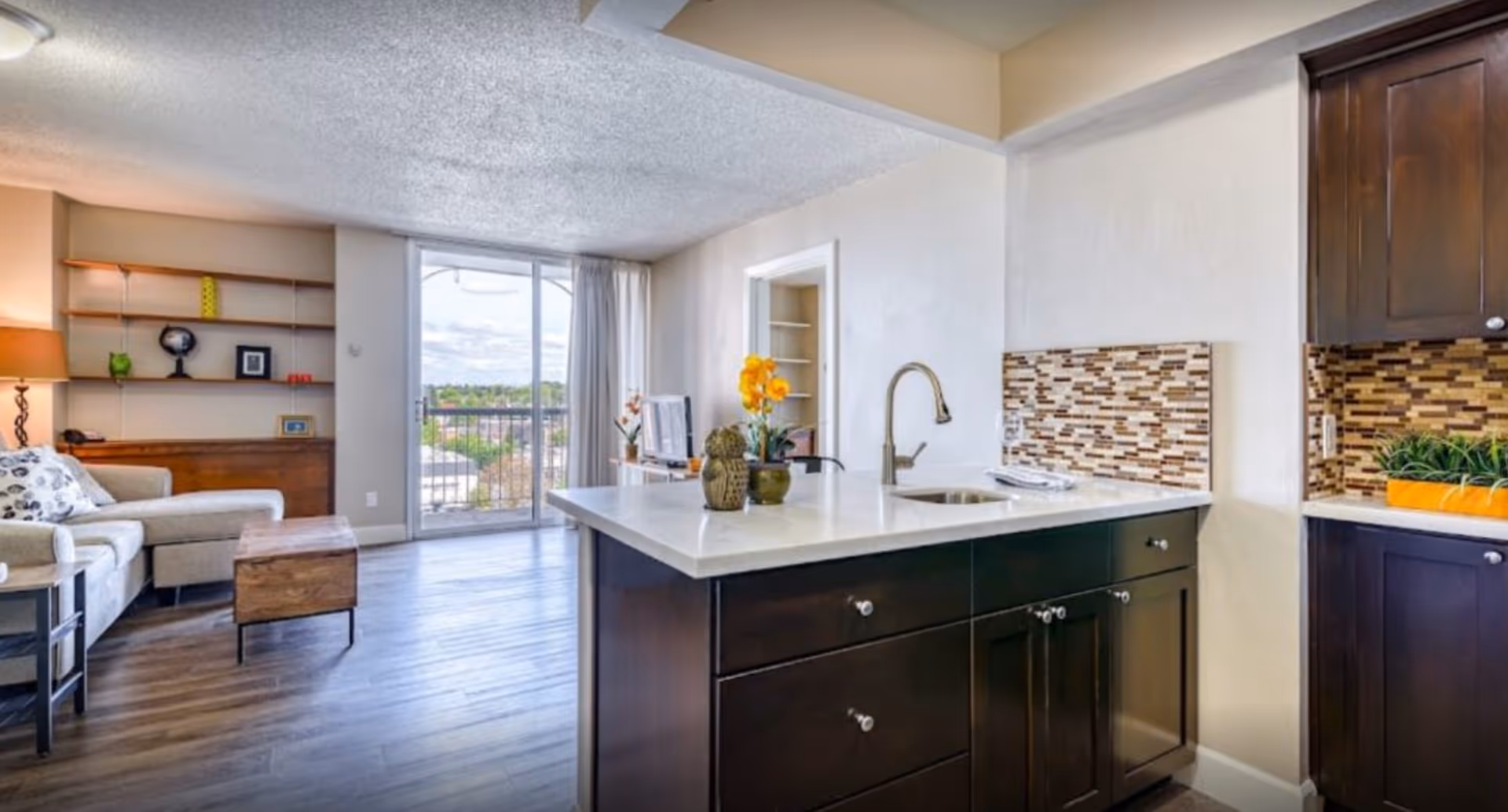 Open-concept apartment living area with a kitchen island in the foreground, seating area to the left, and a sliding glass door to a balcony.