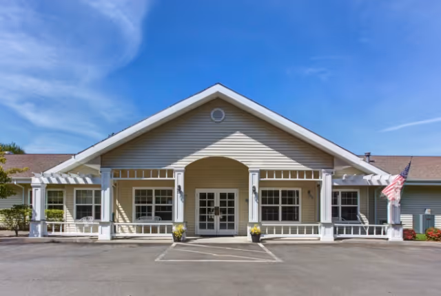 Front exterior view of a single-story building with a peaked roof, white columns, and a covered entrance. There are two white benches on either side of the entrance, potted plants near the door, and an American flag on the right side. The sky is clear and blue.