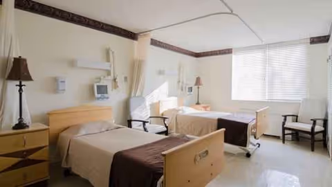 Two beds with beige linens and seating in a bright shared patient room.