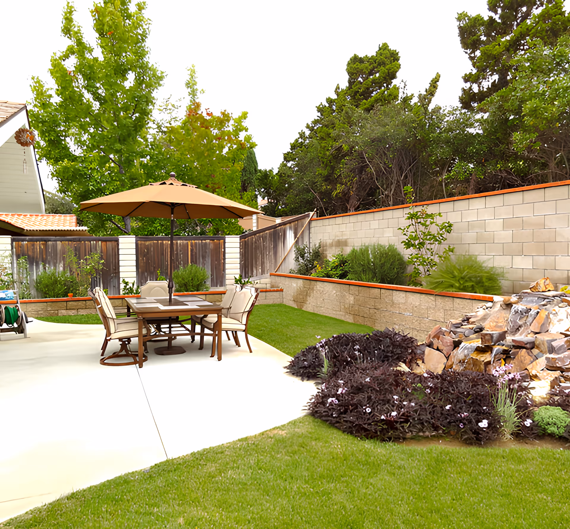Outdoor patio area with a table and four chairs under a large beige umbrella, surrounded by green grass, plants, and trees. There is a stone water feature with rocks and flowing water on the right side, and a wooden fence and brick wall in the background.